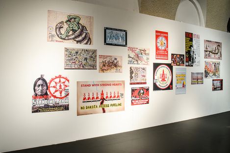 Exhibition wall displaying various protest posters, including messages against the Dakota Access Pipeline. Vibrant illustrations convey activism and resistance.