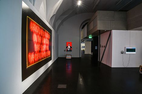 Contemporary gallery interior with a neon sign reading, "ARTISTS NEED TO CREATE ON THE SAME SCALE THAT SOCIETY HAS THE CAPACITY TO DESTROY," conveying urgency.