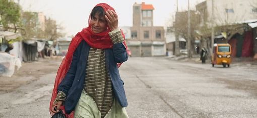 Eine Person mit rotem Kopftuch, blauem Oberteil und gestreiftem Hemd trägt eine schwarze Plastiktüte und geht auf einer leeren Straße in einer Stadt.