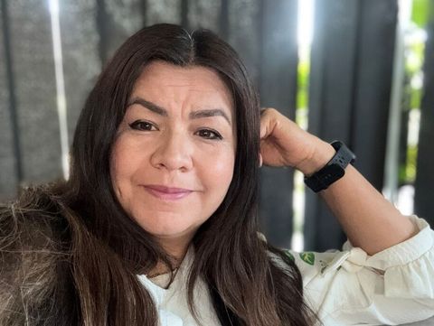 Frau mit langen dunklen Haaren trägt weiße Bluse und schwarzen Armbanduhr sitzt vor schwarzen vertikalen Lamellen.
