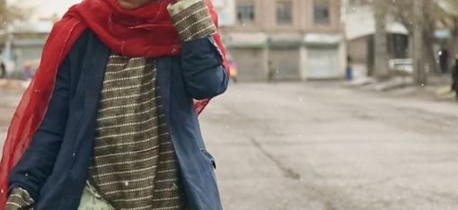 Person mit rotem Schal und blauem Mantel geht auf einer leeren Straße mit Gebäuden und einem gelben Tuk-Tuk im Hintergrund