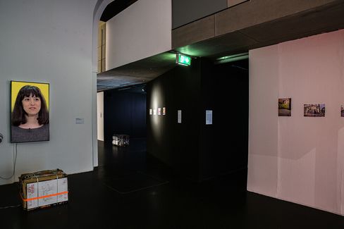 A dimly lit art gallery displays a video portrait of a woman on a wall, with photos on adjacent walls. The atmosphere is modern and contemplative.