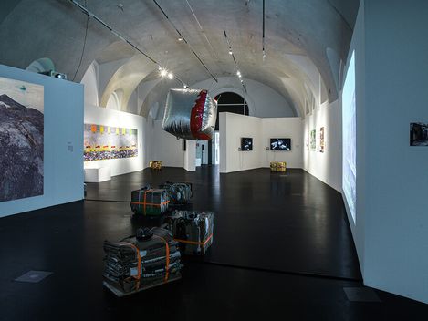 Art gallery with dim lighting and vaulted ceiling features contemporary installations, hanging sculpture, wrapped objects, and diverse wall artworks.