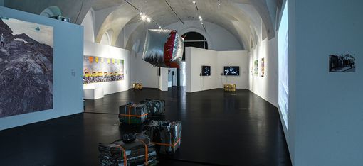 Art gallery with dim lighting and vaulted ceiling features contemporary installations, hanging sculpture, wrapped objects, and diverse wall artworks.