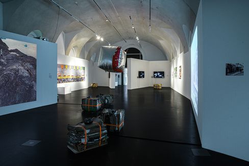 Art gallery with dim lighting and vaulted ceiling features contemporary installations, hanging sculpture, wrapped objects, and diverse wall artworks.