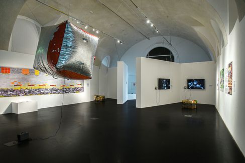 A modern art gallery with a high ceiling features a metallic cube sculpture hanging from above. Walls showcase colorful, abstract artwork and two TVs. The ambiance is contemporary and reflective.