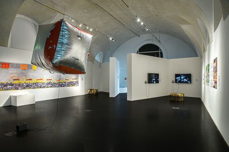 A modern art gallery with a high ceiling features a metallic cube sculpture hanging from above. Walls showcase colorful, abstract artwork and two TVs. The ambiance is contemporary and reflective.