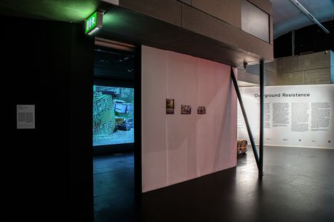 Gallery exhibit titled "Overground Resistance." Dimly lit room with photos on a white wall, a lit emergency exit, and a vibrant protest banner visible inside.