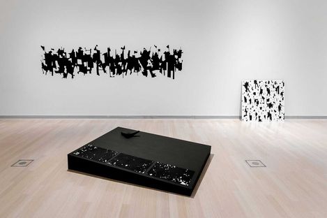 Schwarz-weiße abstrakte Kunstwerke an weißer Wand und auf schwarzem Podest in einem hellen Ausstellungsraum.