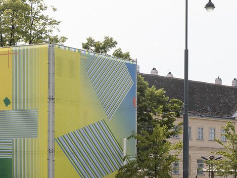 Large rectangular object with colorful geometric patterns and the text 'MQ' on a red circle in front of historic buildings.