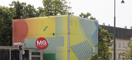 Large rectangular object with colorful geometric patterns and the text 'MQ' on a red circle in front of historic buildings.