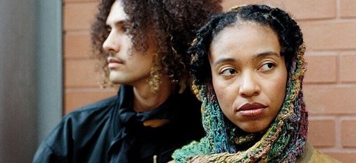 Two people in front of a brick wall, one with curly hair looking sideways, the other with braided hair and colorful scarf looking to the right.