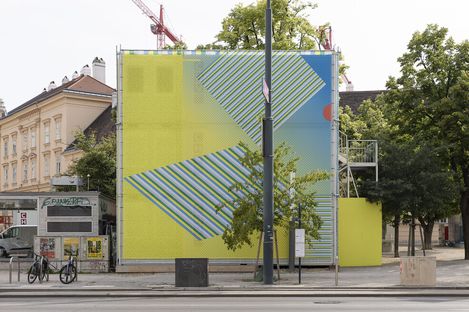Large mural with diagonal stripes and geometric shapes on a building facade, with a streetlight, trees, and bicycles in front.