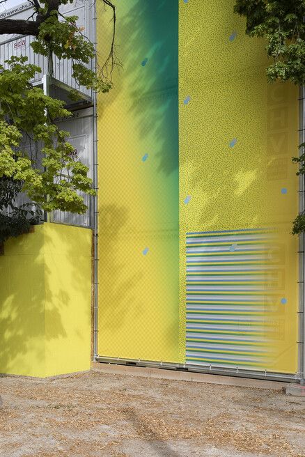 Large wall with a yellow-green gradient, small blue dots, and a section with horizontal stripes, partly shaded by branches.