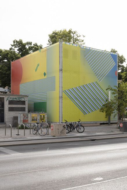 Cube-shaped building with large-scale facade design of geometric patterns and color gradients, with several bicycles and bike racks in front.