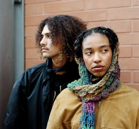Two people in front of a brick wall, one with curly hair looking sideways, the other with braided hair and colorful scarf looking to the right.