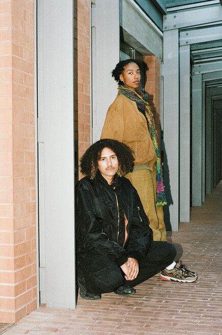 Two people in a corridor, one sitting on the floor wearing a black jacket with curly hair, the other standing in the background wearing a brown jacket and colorful scarf.
