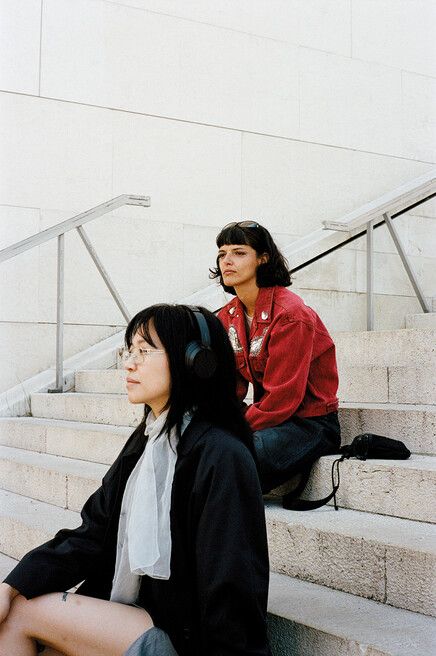 Two women sitting on stairs, one wearing headphones and glasses, the other in a red jacket with sunglasses on her head.