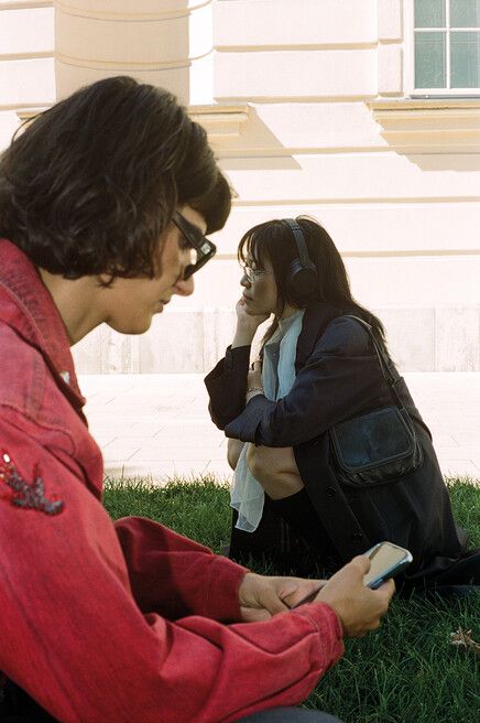 Two people sitting on grass in front of a light-colored wall, one wearing headphones looking thoughtfully to the side, the other holding a smartphone wearing a red jacket.