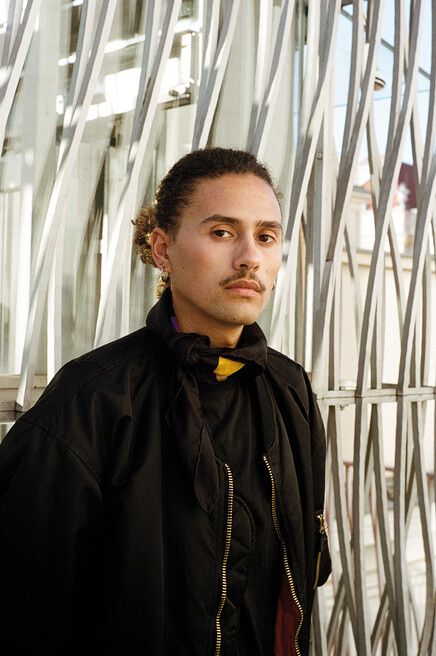 Portrait of a person with tied-back hair, mustache, and earring wearing a black jacket and a black bow tie with a yellow accent standing in front of a lattice of white metal bars.