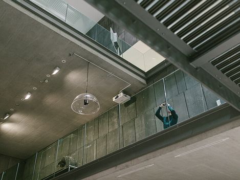 Blick von unten auf moderne Innenarchitektur mit Glasgeländer; eine Person fotografiert.