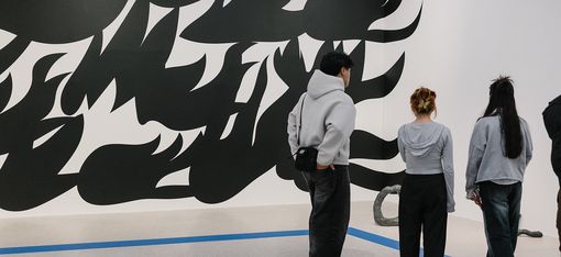 Drei Personen stehen vor einer weißen Wand mit einer großflächigen schwarz-weißen abstrakten Wandmalerei in einem Ausstellungsraum.