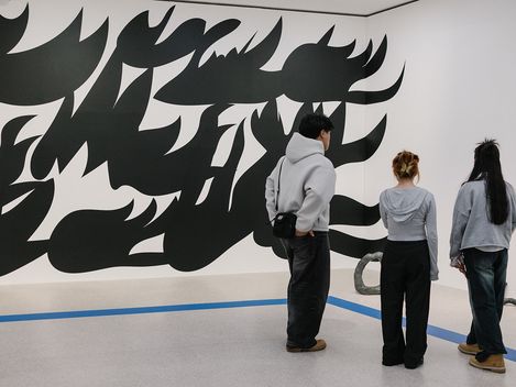 Drei Personen stehen vor einer weißen Wand mit einer großflächigen schwarz-weißen abstrakten Wandmalerei in einem Ausstellungsraum.