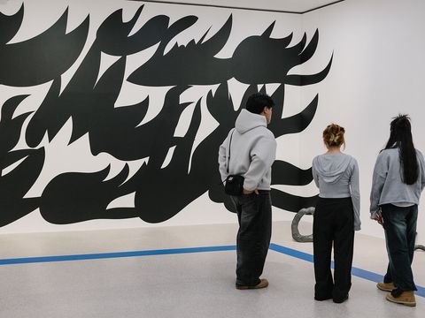 Drei Personen stehen vor einer weißen Wand mit einer großflächigen schwarz-weißen abstrakten Wandmalerei in einem Ausstellungsraum.