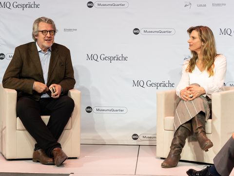 Zwei Personen sitzen auf weißen Sesseln vor einer Wand mit dem Schriftzug "MQ Gespräche" und "MuseumsQuartier", beide tragen formelle Kleidung und sind in einem Gespräch vertieft.
