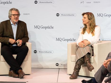 Two people sit on white armchairs in front of a wall with the text "MQ Gespräche" and "MuseumsQuartier", both dressed formally and engaged in conversation.