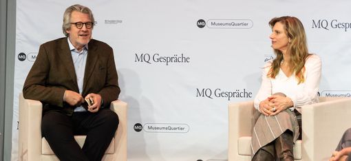 Zwei Personen sitzen auf weißen Sesseln vor einer Wand mit dem Schriftzug "MQ Gespräche" und "MuseumsQuartier", beide tragen formelle Kleidung und sind in einem Gespräch vertieft.
