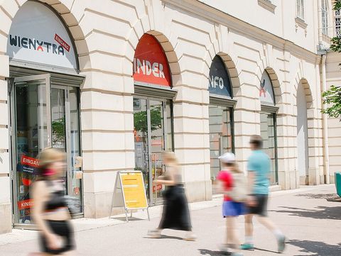 The image shows the exterior of a bright, historic building with several large arched entrances. Above the doors, signs reading “WIENXTRA” and “Kinderinfo” are visible. People, including adults and children, are walking past the building; some appear slightly blurred in motion, conveying a lively, urban atmosphere. On the right side of the image, there are modern, colorful seating or play elements in the outdoor area, along with a tree providing shade. Overall, the image gives the impression of an open, welcoming place for families and children in the heart of the city.