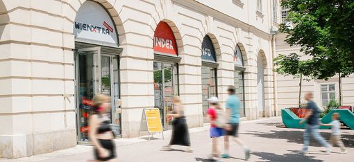 The image shows the exterior of a bright, historic building with several large arched entrances. Above the doors, signs reading “WIENXTRA” and “Kinderinfo” are visible. People, including adults and children, are walking past the building; some appear slightly blurred in motion, conveying a lively, urban atmosphere. On the right side of the image, there are modern, colorful seating or play elements in the outdoor area, along with a tree providing shade. Overall, the image gives the impression of an open, welcoming place for families and children in the heart of the city.