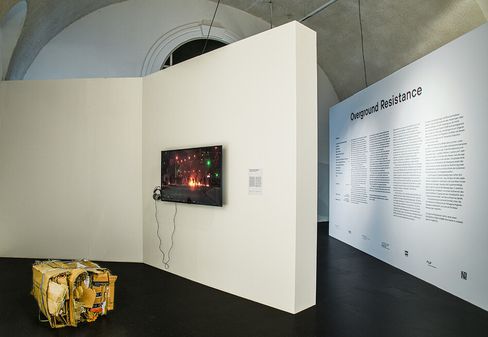 An exhibition space with a white wall displaying a screen with headphones and a large wall text titled 'Overground Resistance', next to a box-shaped object with yellow and brown surfaces.