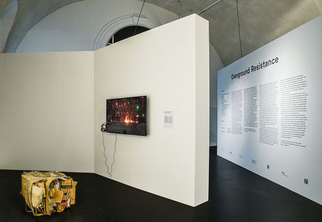 An exhibition space with a white wall displaying a screen with headphones and a large wall text titled 'Overground Resistance', next to a box-shaped object with yellow and brown surfaces.