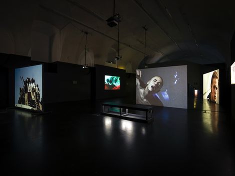 Dark exhibition room with multiple large video projections displaying intense scenes on the walls. A bench is centered under overhead projectors.