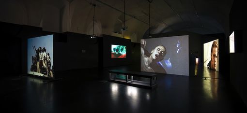 Dark exhibition room with multiple large video projections displaying intense scenes on the walls. A bench is centered under overhead projectors.
