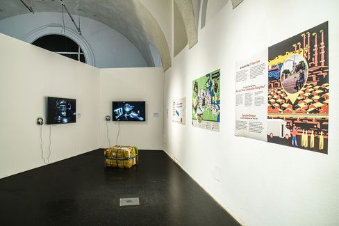 Exhibition room with two monitors on the wall, headphones, a rectangular object on the floor, and several large wall panels with text and graphics.