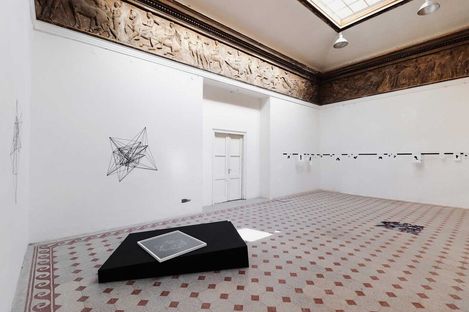 Großer heller Ausstellungsraum mit weiß gestrichenen Wänden, einem kunstvollen Fries unter der Decke und geometrischen Drahtskulpturen an den Wänden und auf einem schwarzen Podest auf dem Boden.
