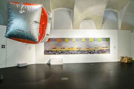 Large silver and red cube floats in front of a wall with a long rectangular artwork and several small objects on the floor.