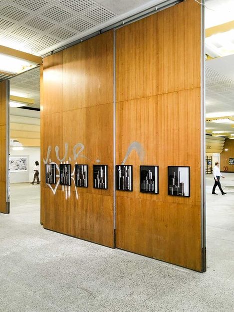 Holzpaneele mit mehreren schwarz-weißen gerahmten Fotografien, die an einer Wand in einem Ausstellungsraum hängen, mit Personen im Hintergrund.