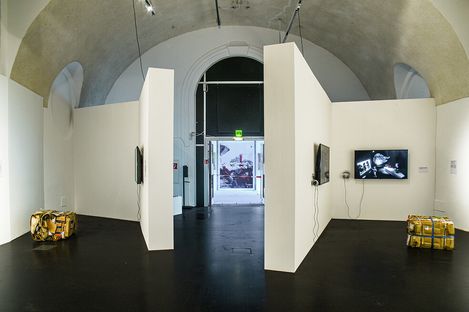 View into an exhibition space with white partition walls, two monitors on the walls, and two cuboid objects on the floor.