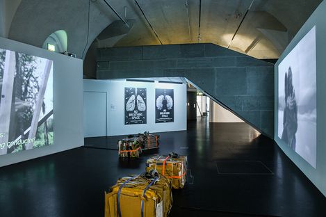 Several yellow, strapped packages lie on the floor of an exhibition space with large wall projections and three posters showing lung motifs on the wall.