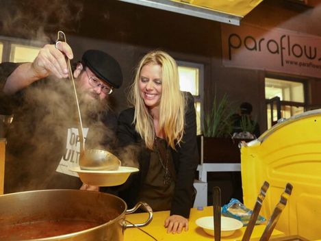 Zwei Personen in einer Küche, eine schöpft Suppe aus einem großen Topf in eine Schüssel