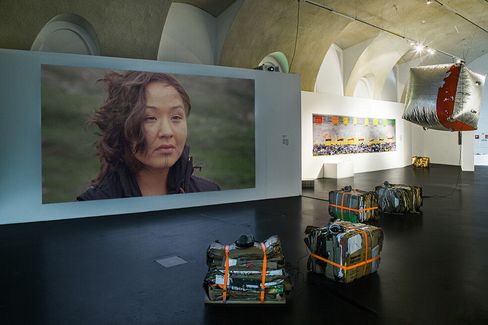 An art gallery displays a large portrait of a contemplative person on a wall. The room also has abstract sculptures, including hanging and boxed pieces, under arched ceilings.