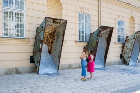Zwei Frauen stehen vor drei großen, schräg an einer hellen Fassade angelehnten Metallcontainern auf einem gepflasterten Platz.