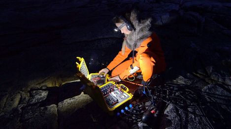 Person in orange protective clothing with headlamp and headphones operating an open yellow technical device on rocky ground in darkness