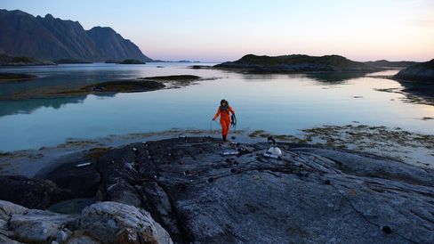 Person in orange protective suit on rocky shore with scattered technical equipment by calm water and mountains in the background