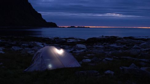 Tent with interior light on rocky ground by the sea at dusk with cloudy sky