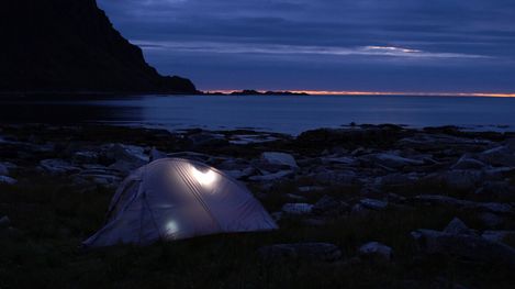 Tent with interior light on rocky ground by the sea at dusk with cloudy sky
